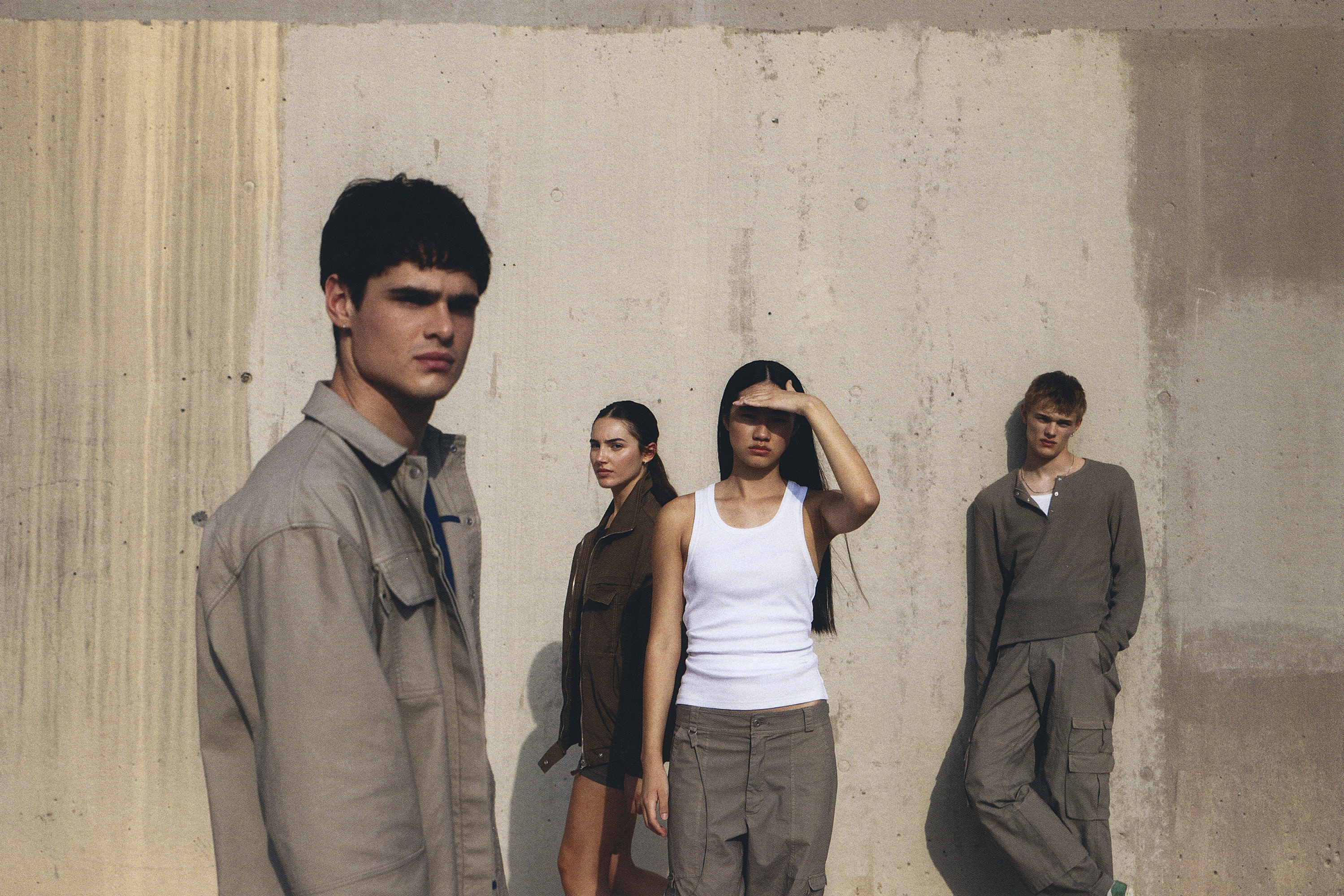 Group of four fashion models in neutral-toned casualwear posing against a concrete wall in natural sunlight.