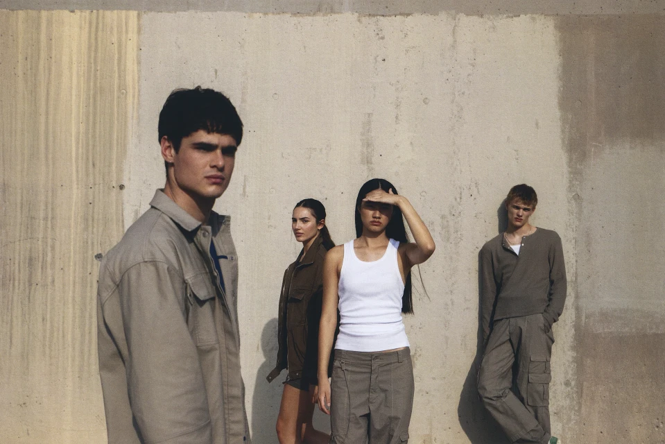 Group of four fashion models in neutral-toned casualwear posing against a concrete wall in natural sunlight.
