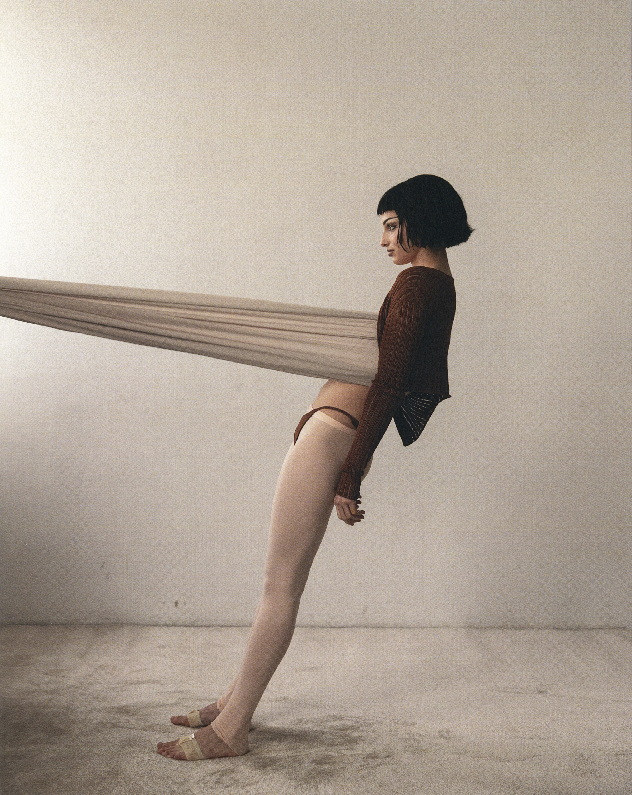 A woman with short dark hair leans backward, supported by a stretched fabric band around her torso in a studio setting.