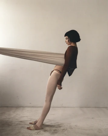 A woman with short dark hair leans backward, supported by a stretched fabric band around her torso in a studio setting.