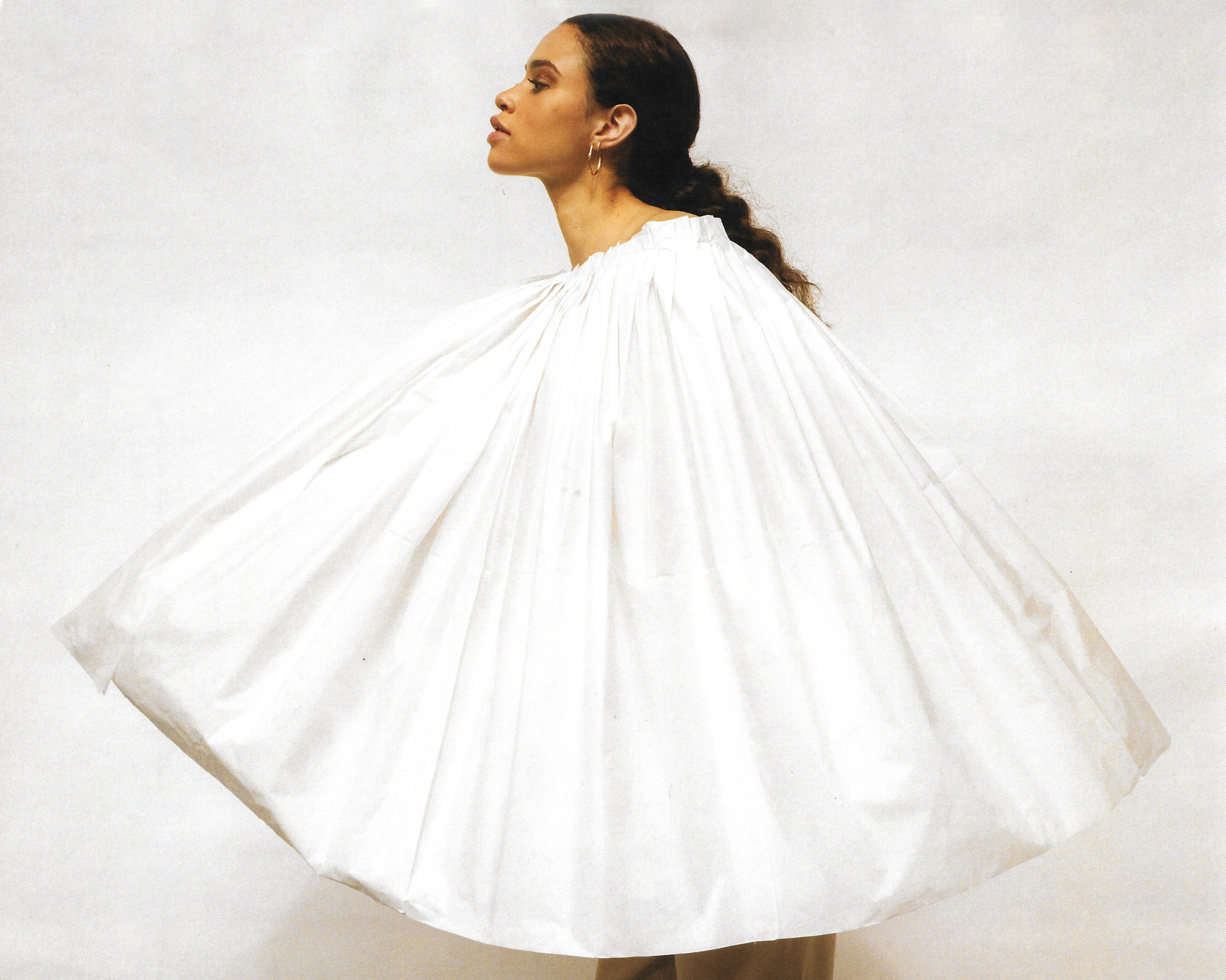 Woman in a flowing white garment stands in a photo studio. The garment has a gathered neckline. Her skin tone is warm and neutral.