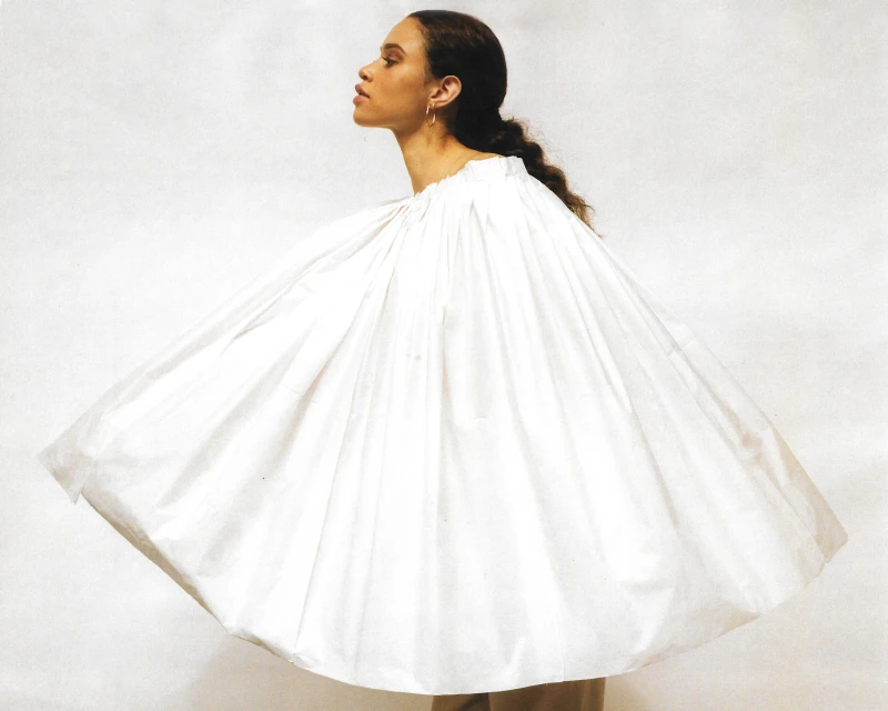 Woman in a flowing white garment stands in a photo studio. The garment has a gathered neckline. Her skin tone is warm and neutral.