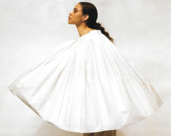 Woman in a flowing white garment stands in a photo studio. The garment has a gathered neckline. Her skin tone is warm and neutral.