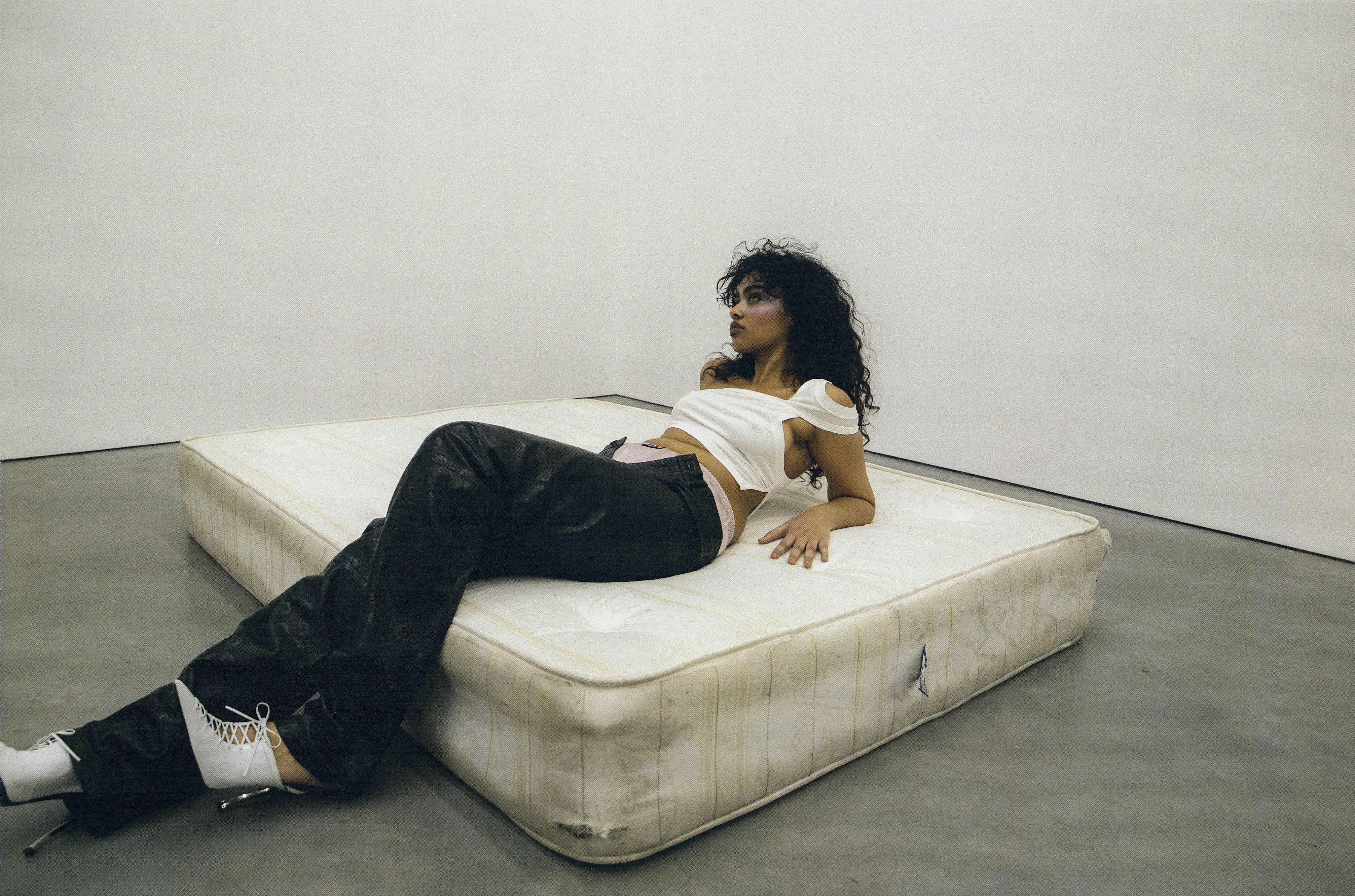 Brunette woman in black pants and white top reclines on mattress in minimalist studio setting, illuminated by diffused lighting.