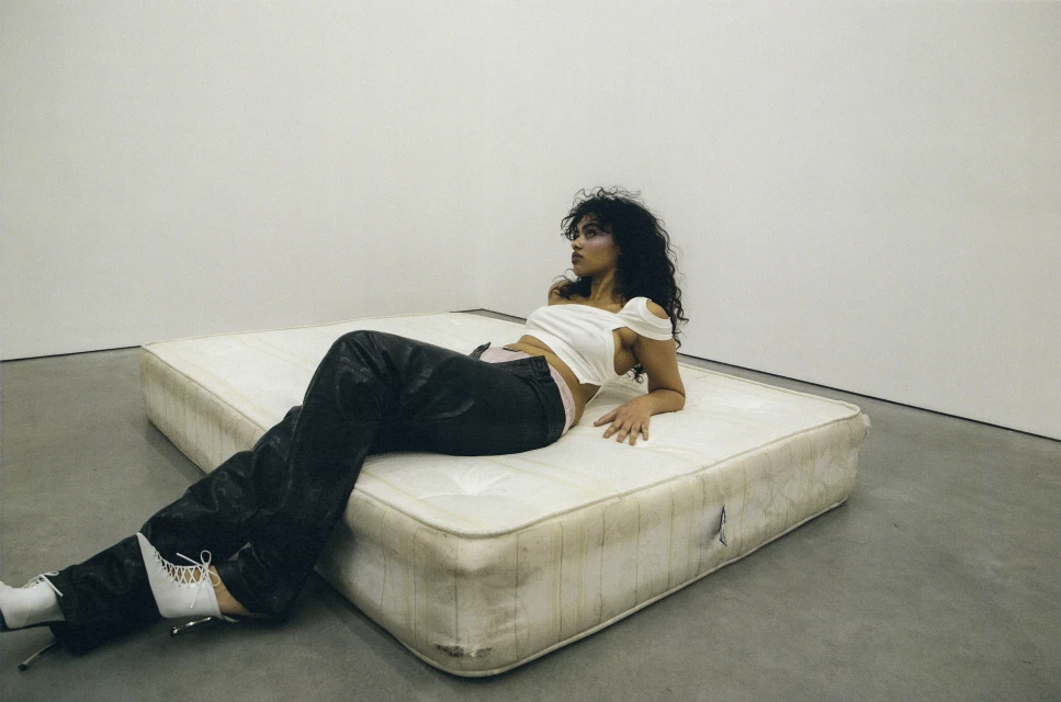 Brunette woman in black pants and white top reclines on mattress in minimalist studio setting, illuminated by diffused lighting.