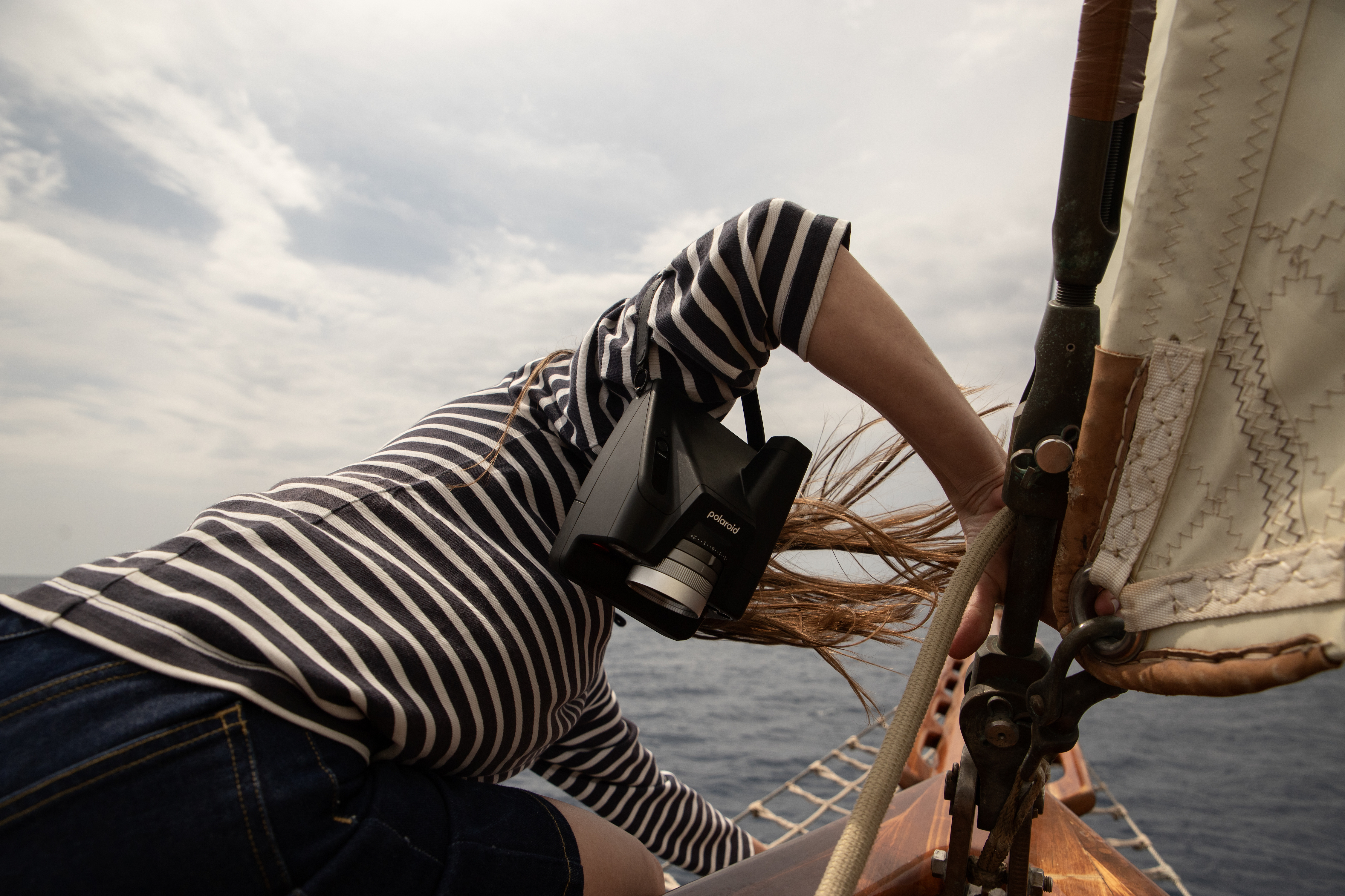 Woman with vintage Polaroid camera on a sailboat, wearing a striped shirt and jeans, with ocean and sky background. Travel and photography.