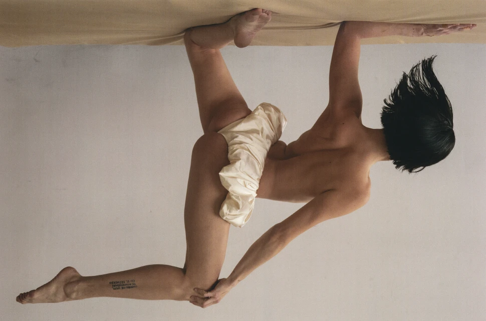 Woman with short dark hair poses, in an upside down shot, gripping a light brown surface and displaying a tattoo on her lower leg.
