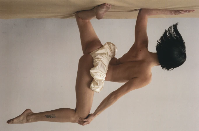 Woman with short dark hair poses, in an upside down shot, gripping a light brown surface and displaying a tattoo on her lower leg.