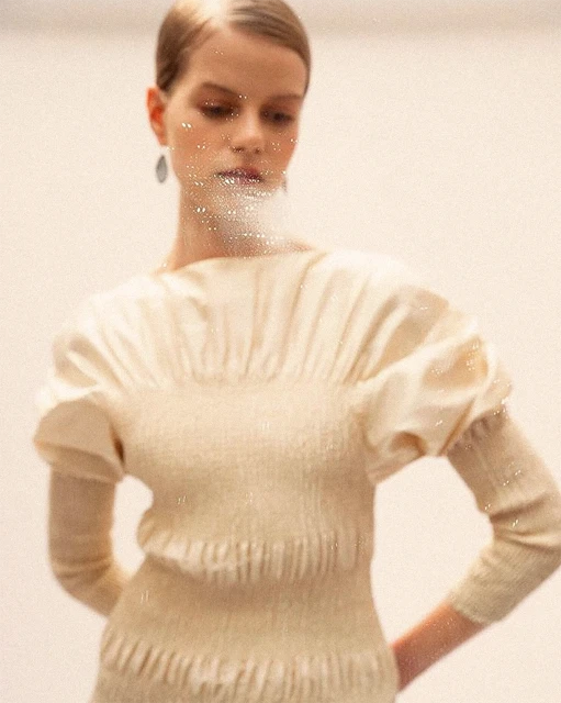 Portrait of a fashion model wearing a beige sweater with puff sleeves. The model has shiny makeup and is photographed against a white background.