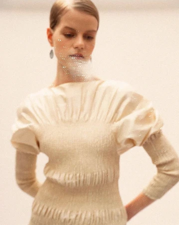 Portrait of a fashion model wearing a beige sweater with puff sleeves. The model has shiny makeup and is photographed against a white background.