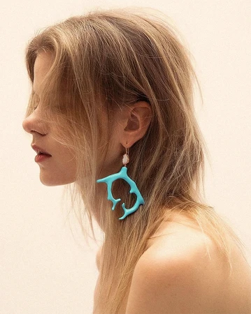 Close-up of a fair-skinned woman in profile wearing a turquoise branch earring and fair blonde hair against a white background.