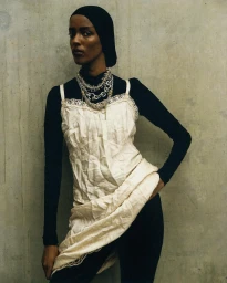 Model in cream slip dress over black top wearing layered silver necklaces against a concrete wall