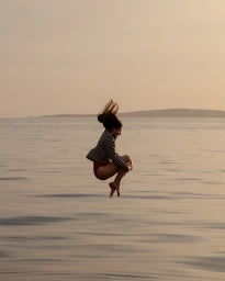 A woman captured mid-air jumping into the ocean at sunset, wearing a striped top and red bottoms.