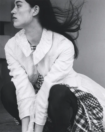 Black and white portrait of a woman crouching outdoors with dark hair blowing in the wind wearing a quilted jacket