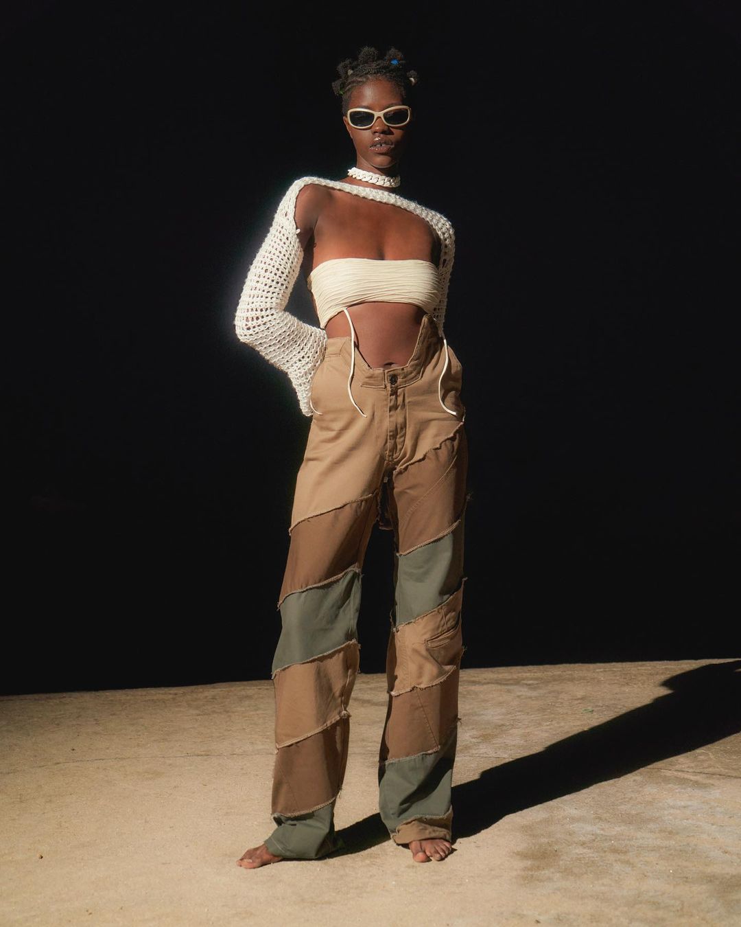 Full-length portrait of a young Black female model in a cream-colored top and patchwork pants against a dark background in a fashion shot.