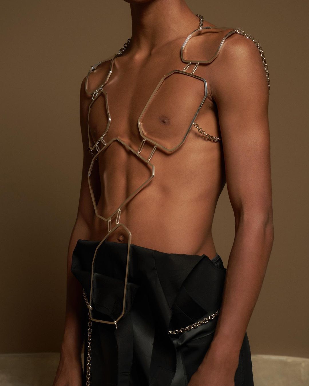 Torso shot of a shirtless male model wearing an avant-garde body chain with a blurred tan background, emphasizing fashion and style.