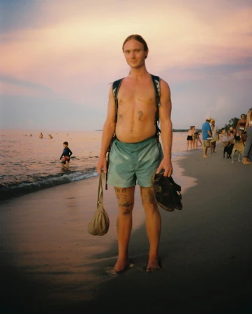 Man standing on a sandy beach holding sandals and a small bag, wearing blue swim trunks and a backpack at sunset