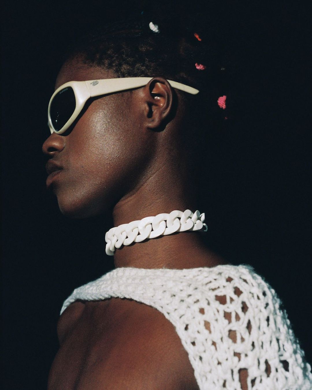 Portrait of an African American person with stylish sunglasses and a chunky white chain necklace, against a dark, shadowy background.