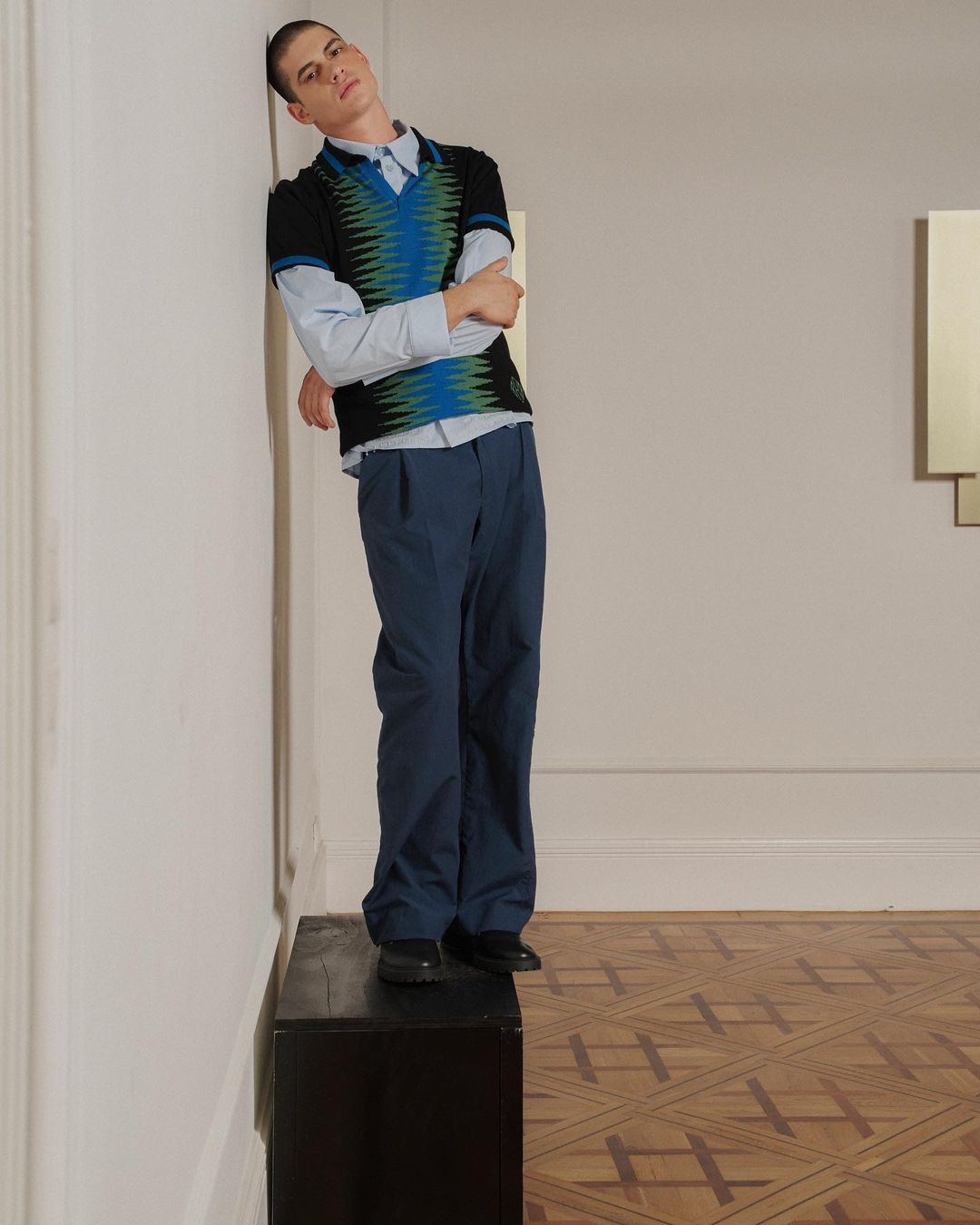 Stylish adult male model poses indoors wearing a black and blue patterned vest with blue trousers in a fashion editorial shot.