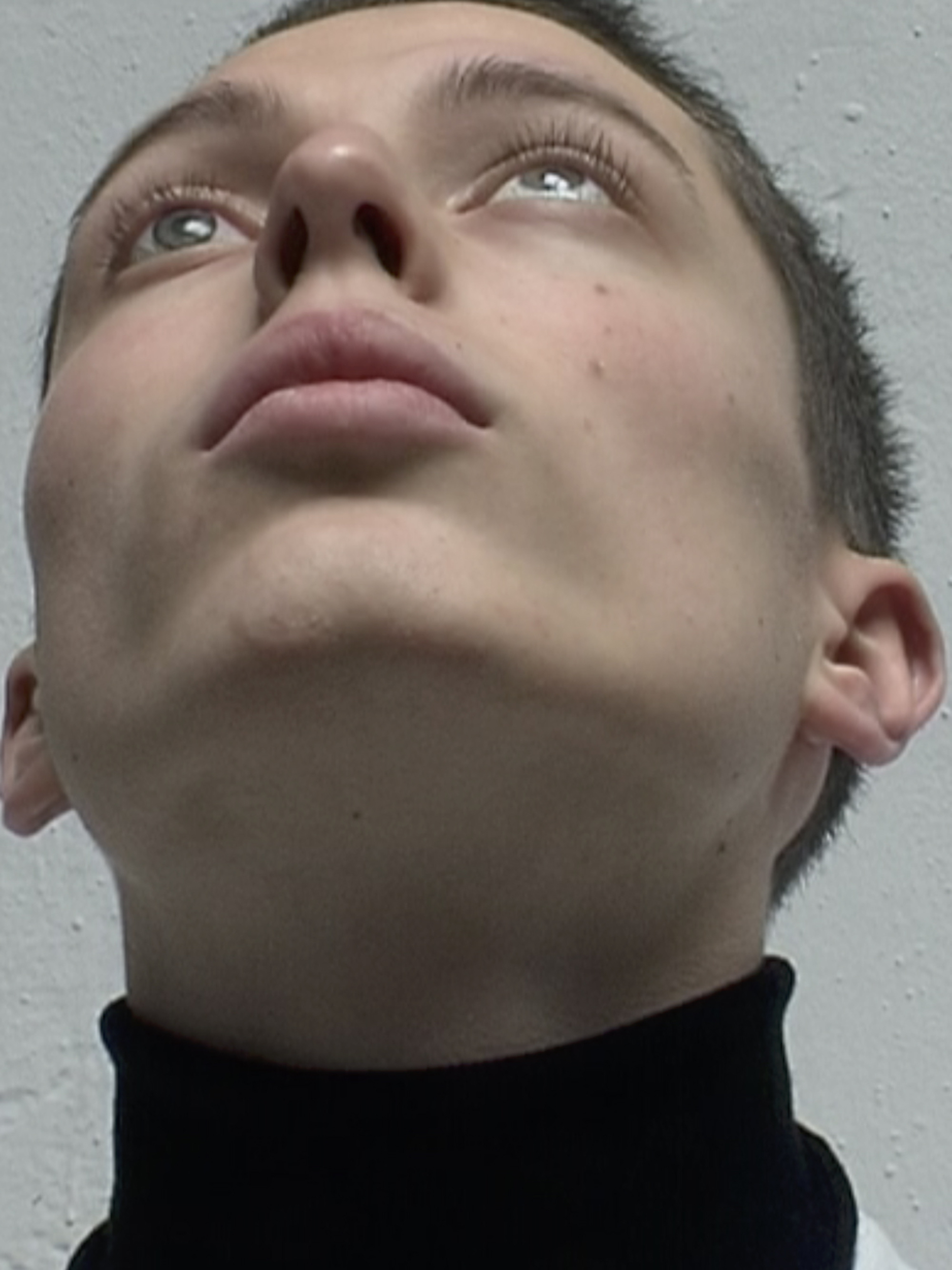 Close-up portrait of a young adult with blue eyes looking upwards against a textured white wall, wearing a black turtleneck top.