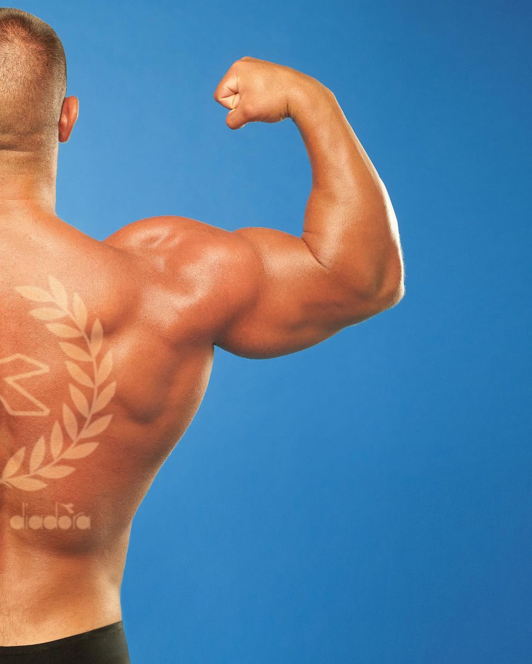 Muscular adult male flexing bicep, back view, with design and Diadora logo on back; studio shot against a vibrant blue background.