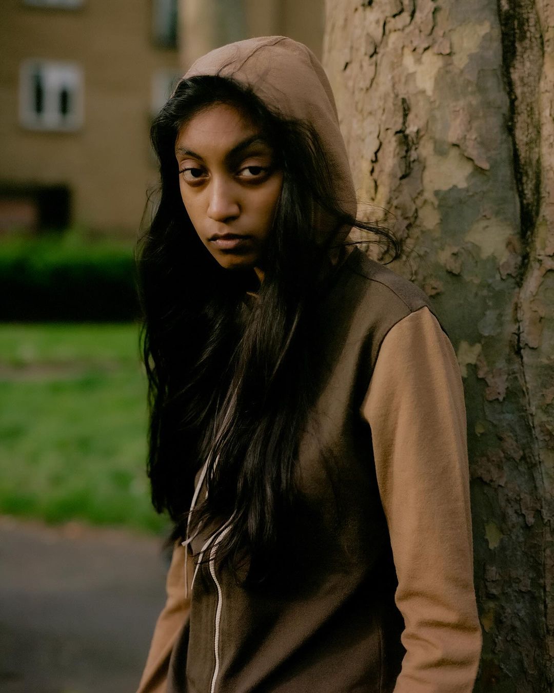 Teenage girl in a hoodie stands against a tree in an urban environment, looking toward the viewer with a neutral expression.