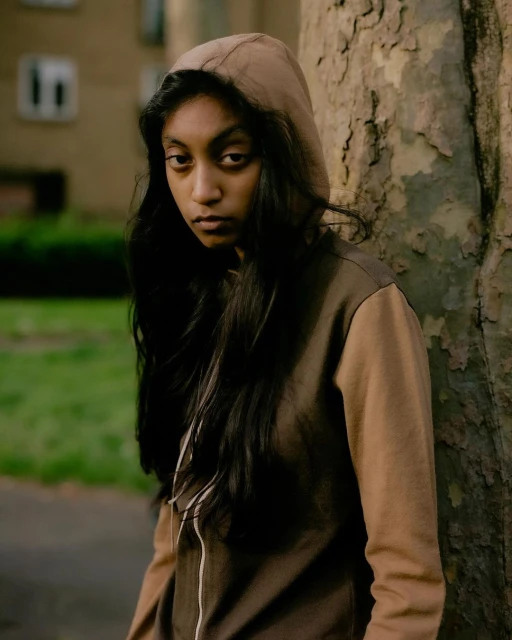 Teenage girl in a hoodie stands against a tree in an urban environment, looking toward the viewer with a neutral expression.