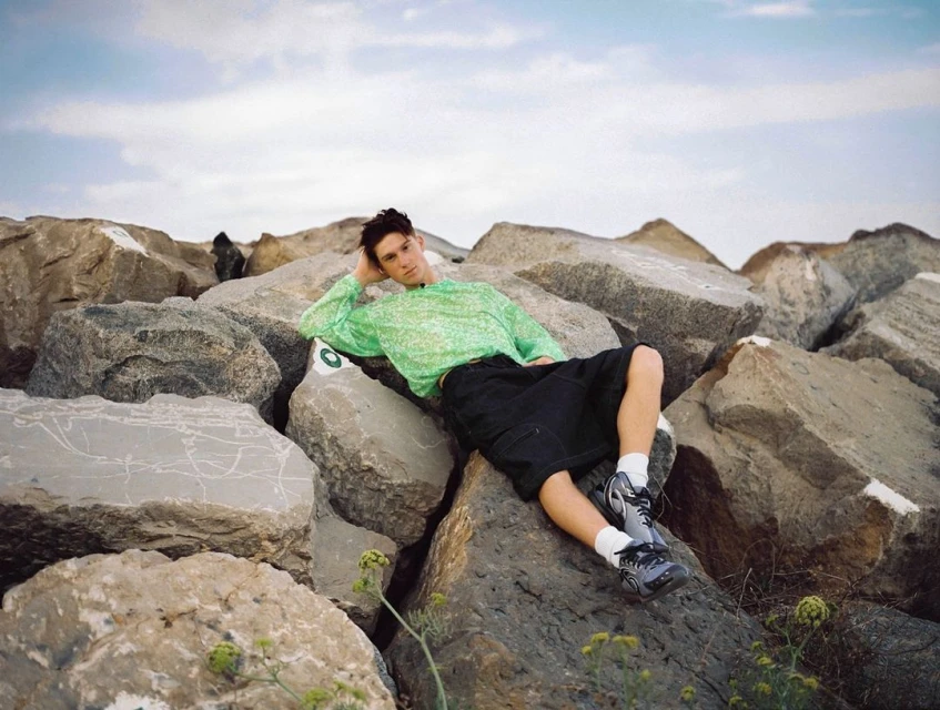A young man with dark hair rests on large rocks near the sea, wearing a green patterned top and black shorts in an outdoor setting.