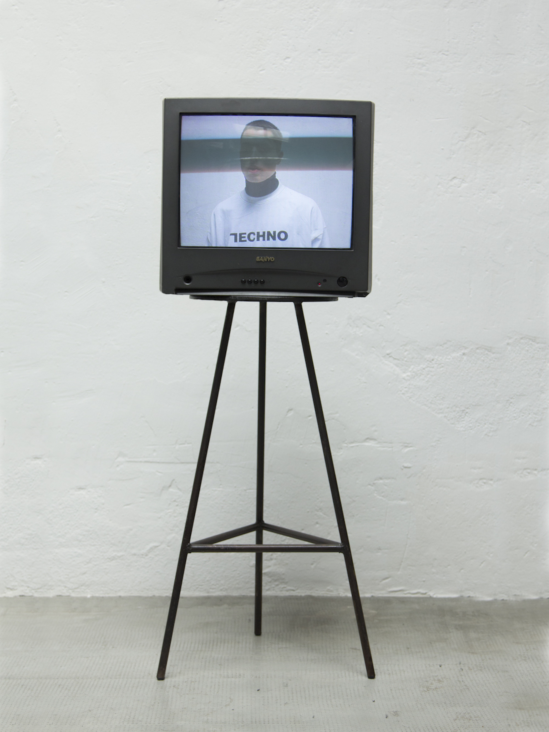 Vintage television on a metal stand displays a person wearing a white shirt with 'Techno' text in a minimalist setting.