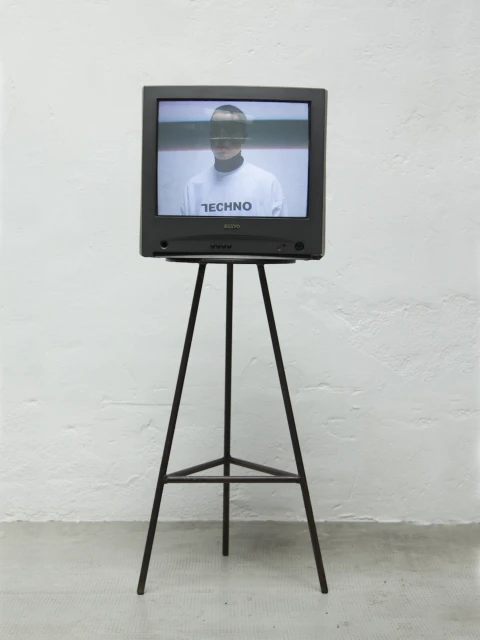 Vintage television on a metal stand displays a person wearing a white shirt with 'Techno' text in a minimalist setting.
