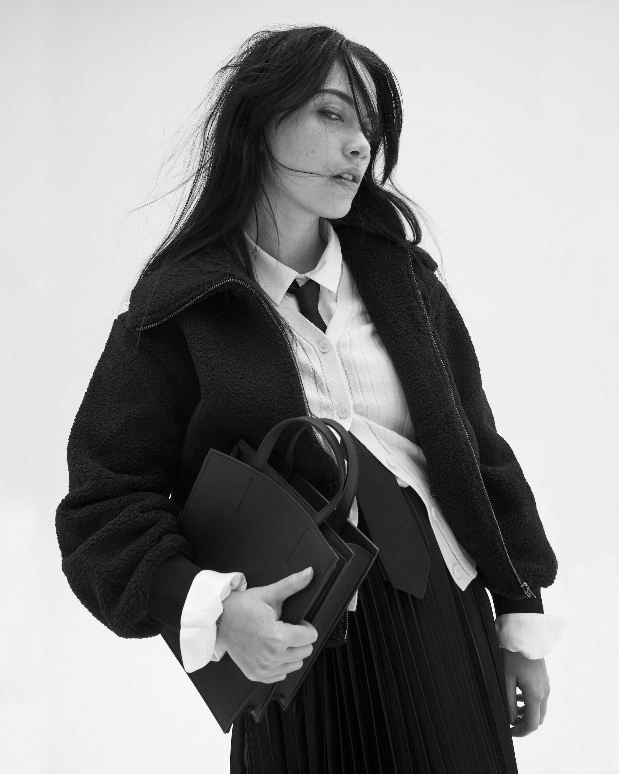 A stylish young adult woman in a black textured jacket, white shirt, and pleated skirt holds a black tote bag against a bright white background.