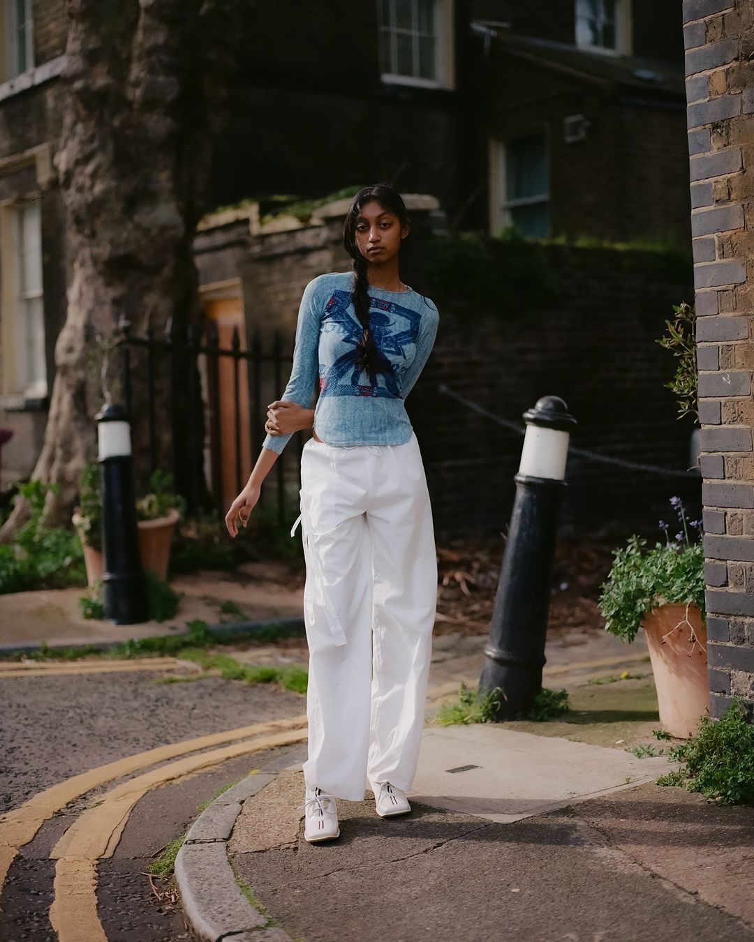 Young woman with long braid poses on urban street in a light blue printed long-sleeved shirt and white cargo pants with white sneakers.