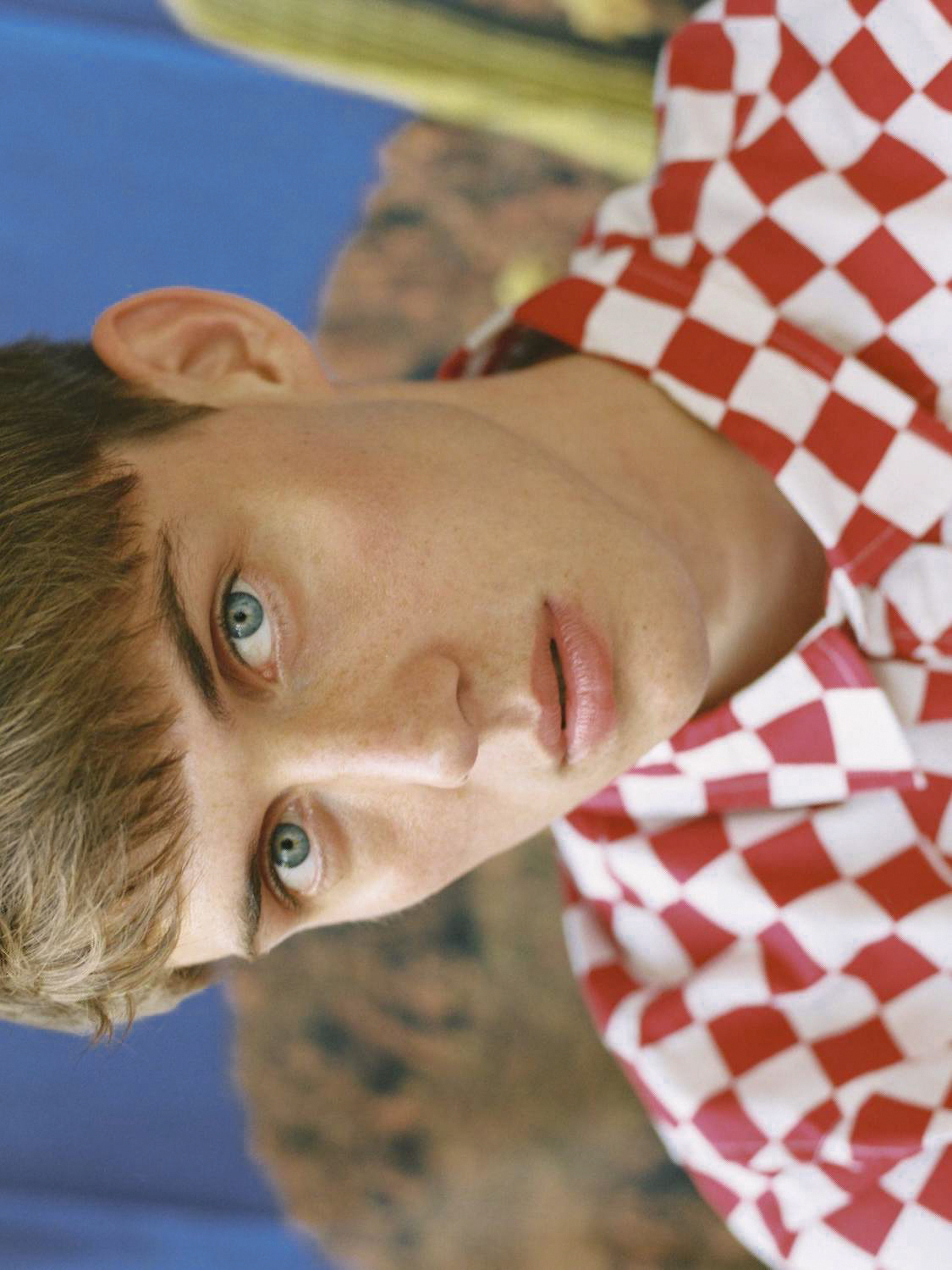 Close-up of a young man with blue eyes and light brown hair wearing a red and white checkered shirt against a blue background.