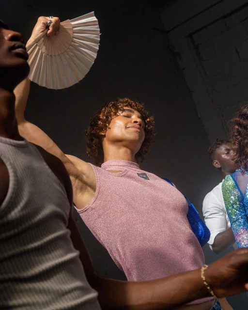 Three stylish people pose indoors in fashionable attire. One person holds a hand fan. The scene has soft lighting, creating a fashionable setting.