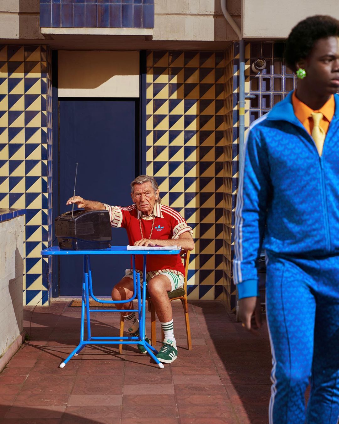 Two men wearing Adidas tracksuits, one seated at a table with a retro radio, the other standing, with an outdoor tiled backdrop.