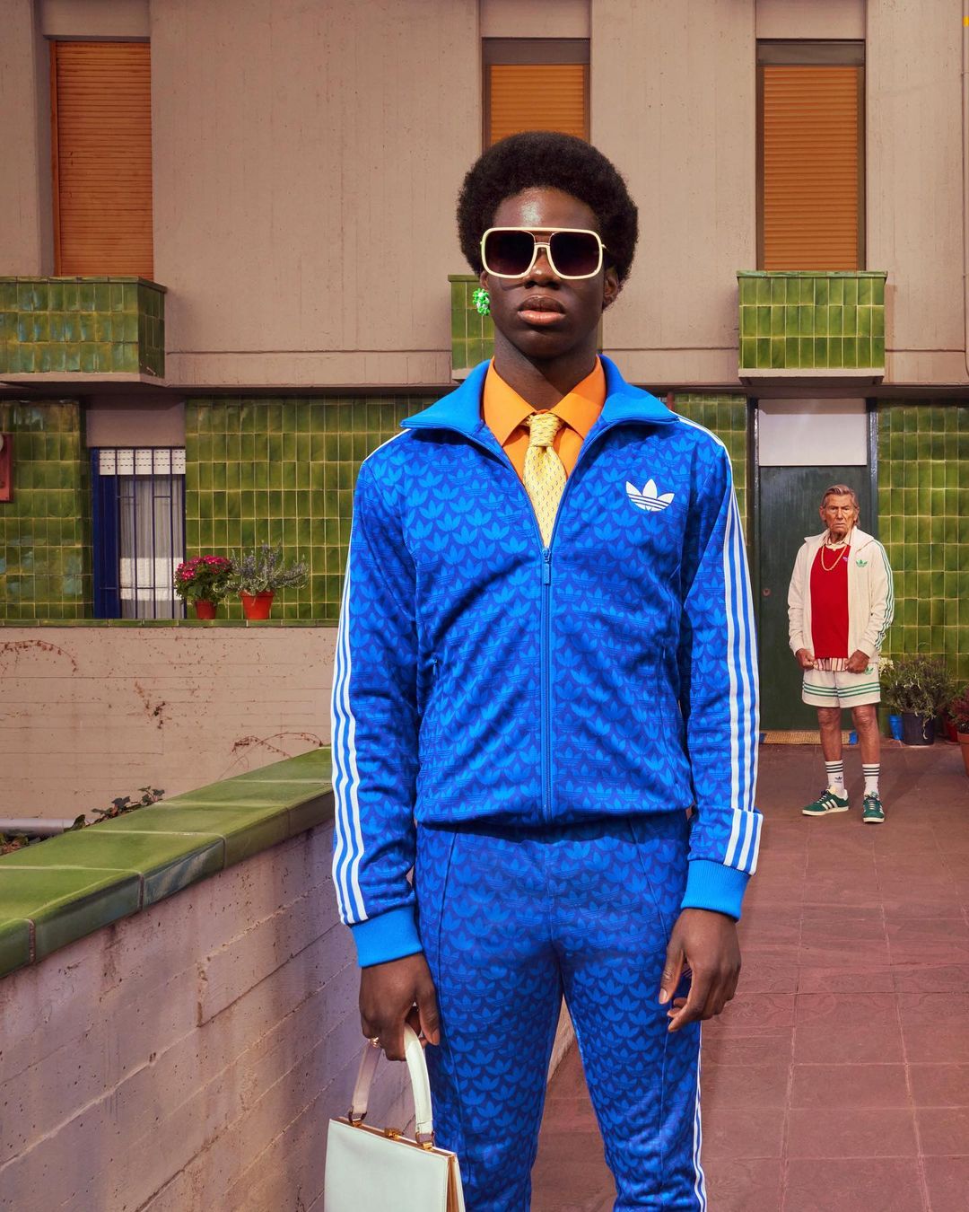A stylish black man in a blue tracksuit poses outdoors with a handbag, while a second man stands in the background, creating a unique fashion statement.