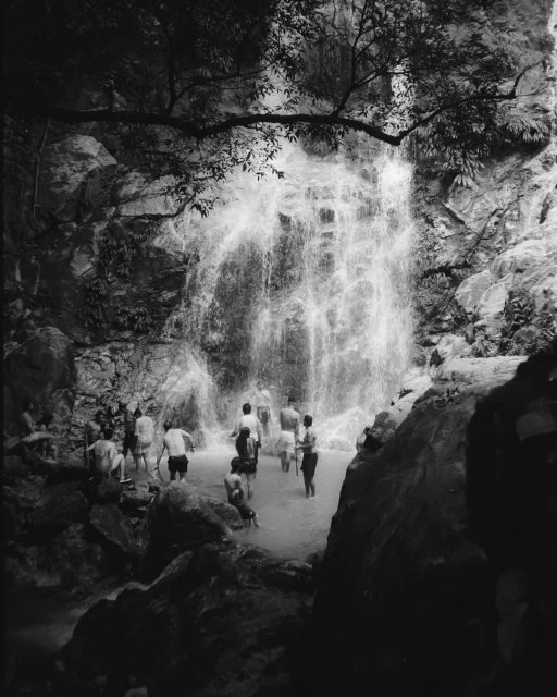 Black and white photograph of a group of people bathing in a pool beneath a waterfall, surrounded by rocks and trees.