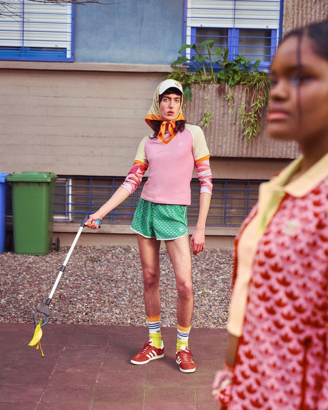 A person in vintage athletic wear uses a trash picker to grab a banana peel, with another person blurred in the foreground.