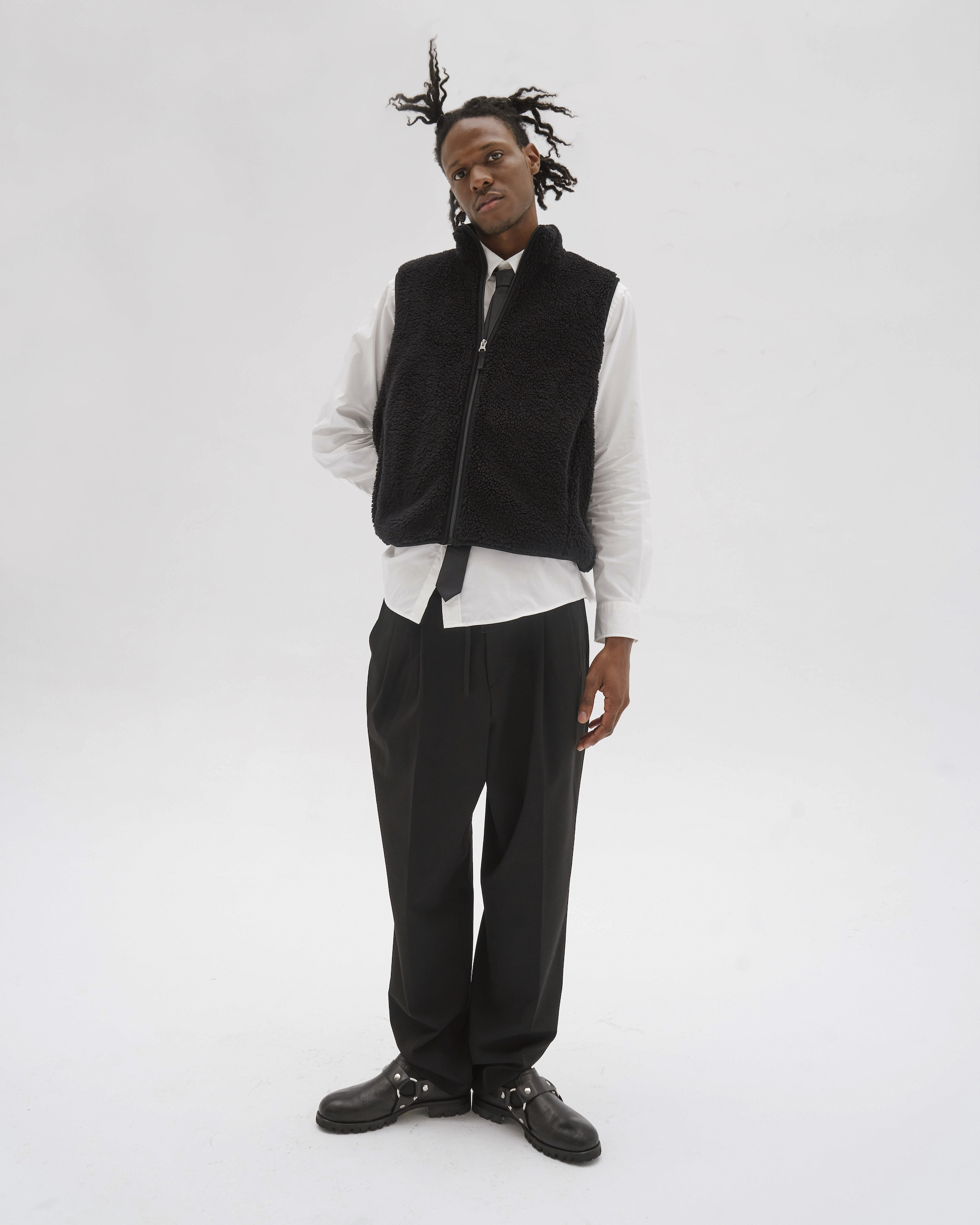 Man in a black vest and white shirt poses in front of a white background. He wears dark pants and shiny black shoes with metal hardware.