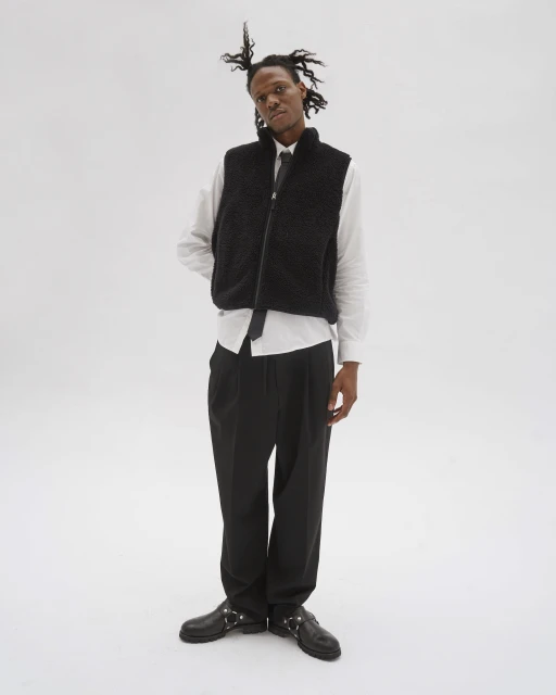 Man in a black vest and white shirt poses in front of a white background. He wears dark pants and shiny black shoes with metal hardware.