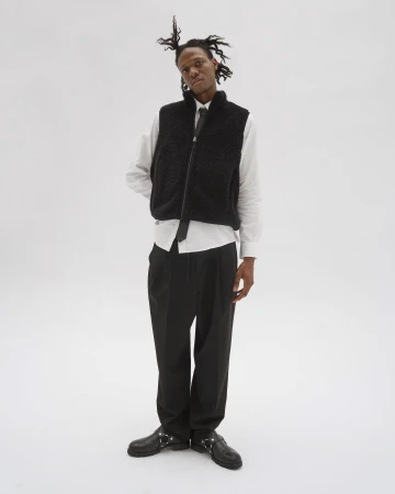 Man in a black vest and white shirt poses in front of a white background. He wears dark pants and shiny black shoes with metal hardware.