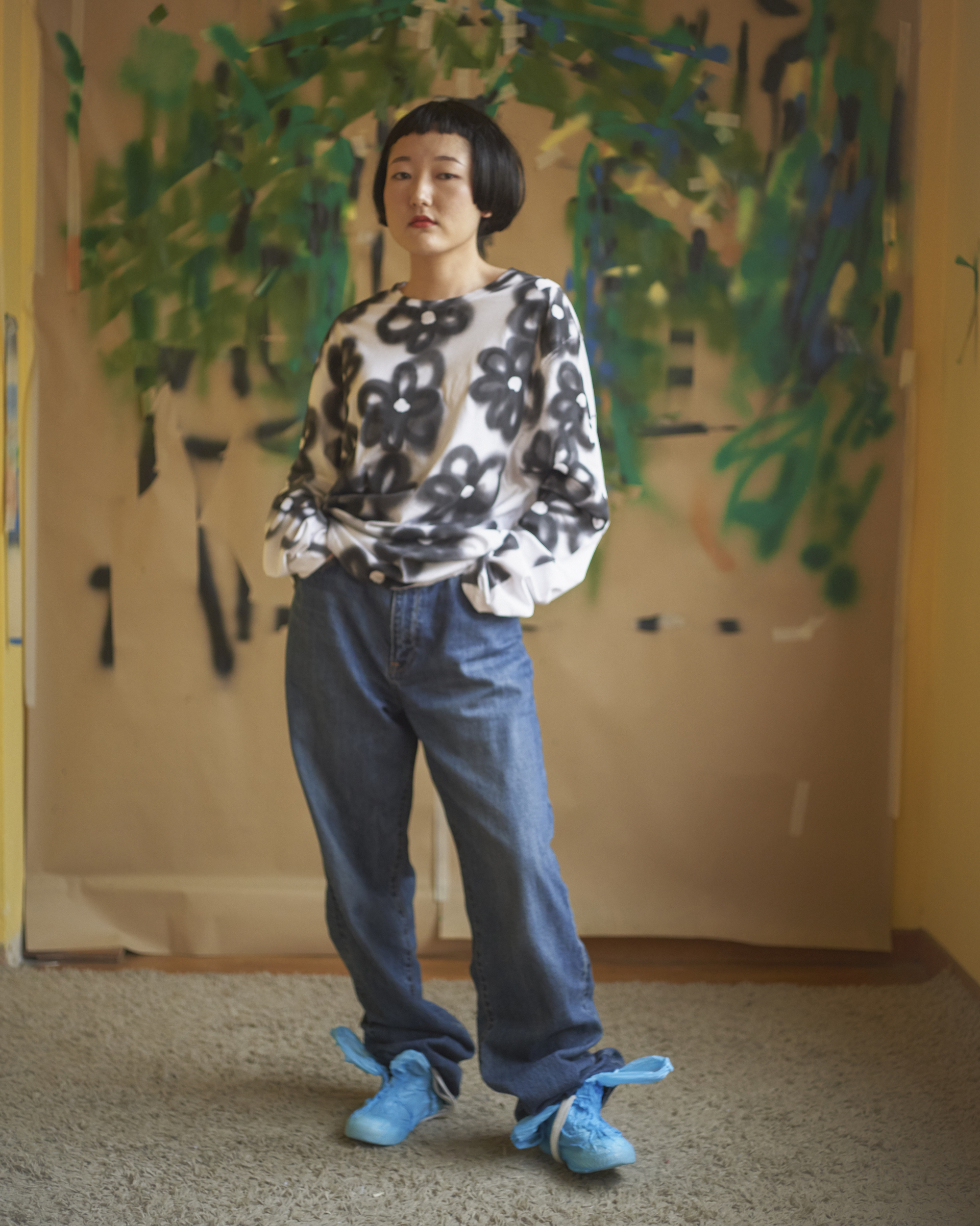 Portrait of a young adult with short dark hair wearing a patterned shirt and jeans, standing in front of a painted backdrop indoors.