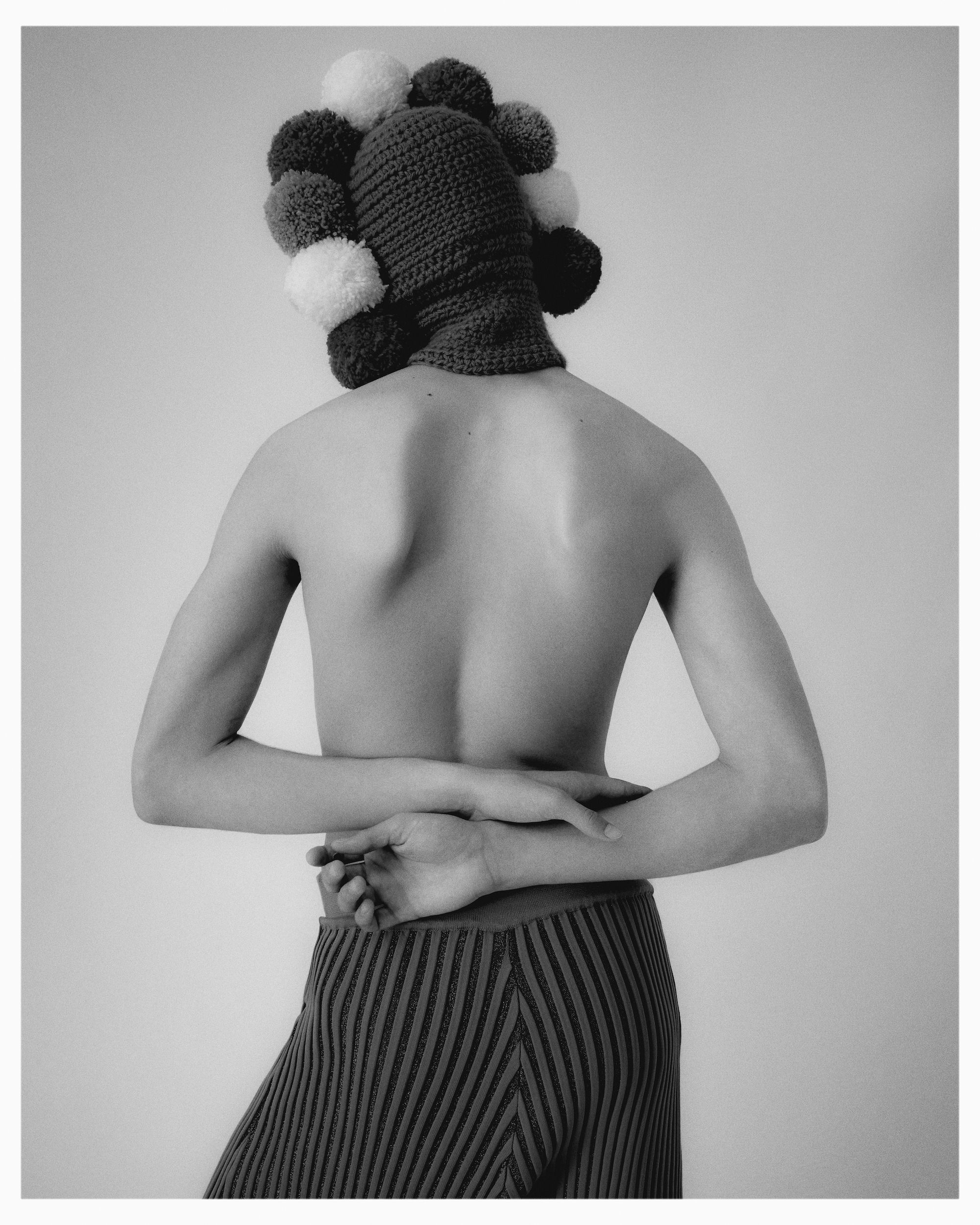 Back view of a slender adult wearing striped pants and a colorful pompom hat, against a solid white backdrop in a studio setting.