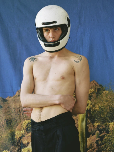 A shirtless man wearing a white motorcycle helmet poses in front of a backdrop with a desert landscape and a blue sky.