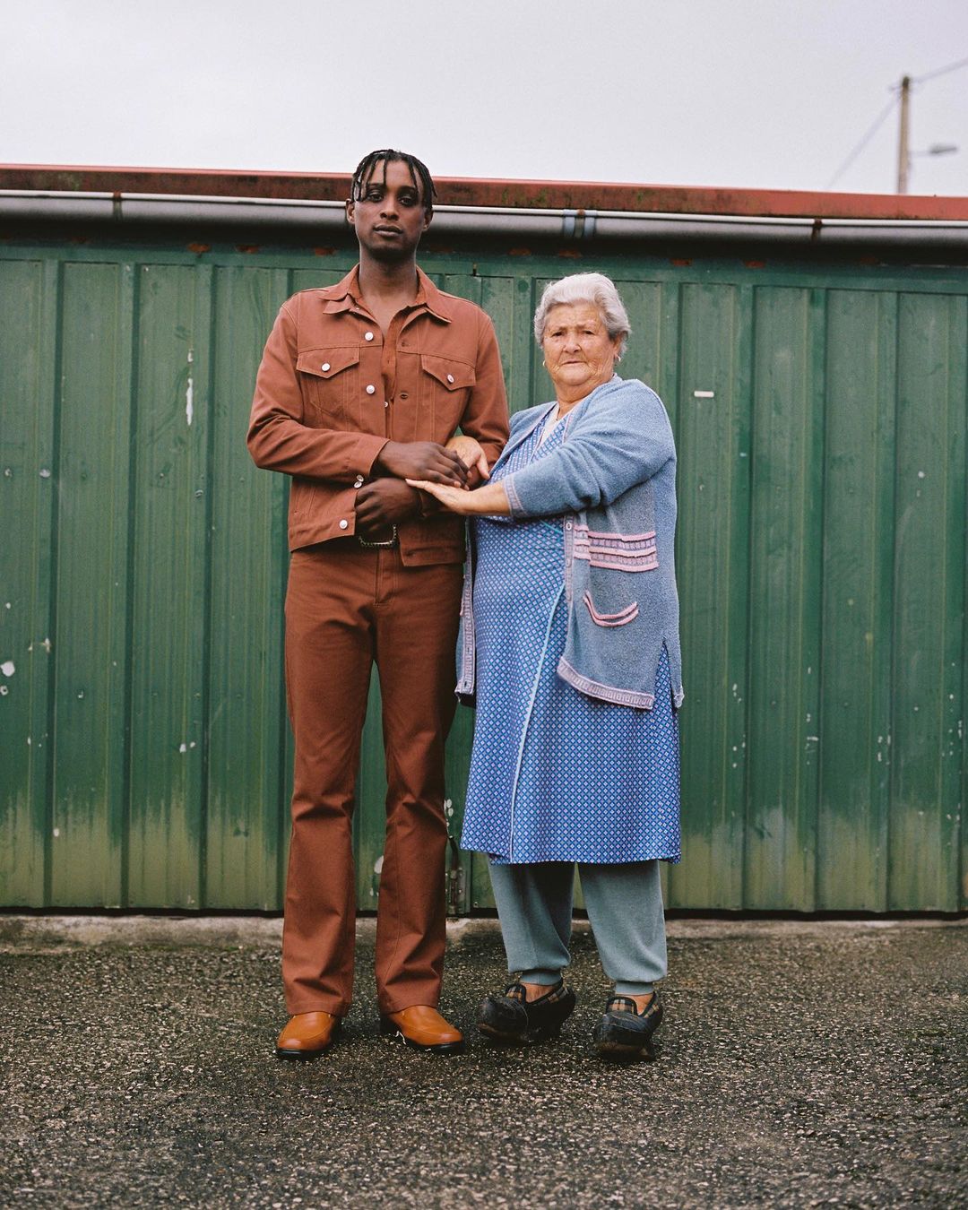 Elderly woman with fair skin and blue clothing poses arm-in-arm with a young Black male wearing brown jacket and pants, standing outdoors.