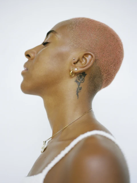 Profile view of a Black female with faded coral hair, ear piercings, a neck tattoo, and wearing a spaghetti-strap top against a white backdrop.