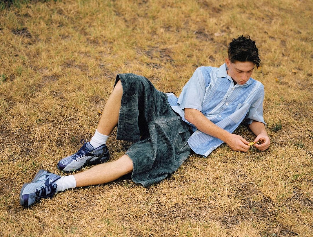 Man lying on grass wearing blue shirt, dark shorts, white socks, and sneakers in an outdoor setting with natural lighting.