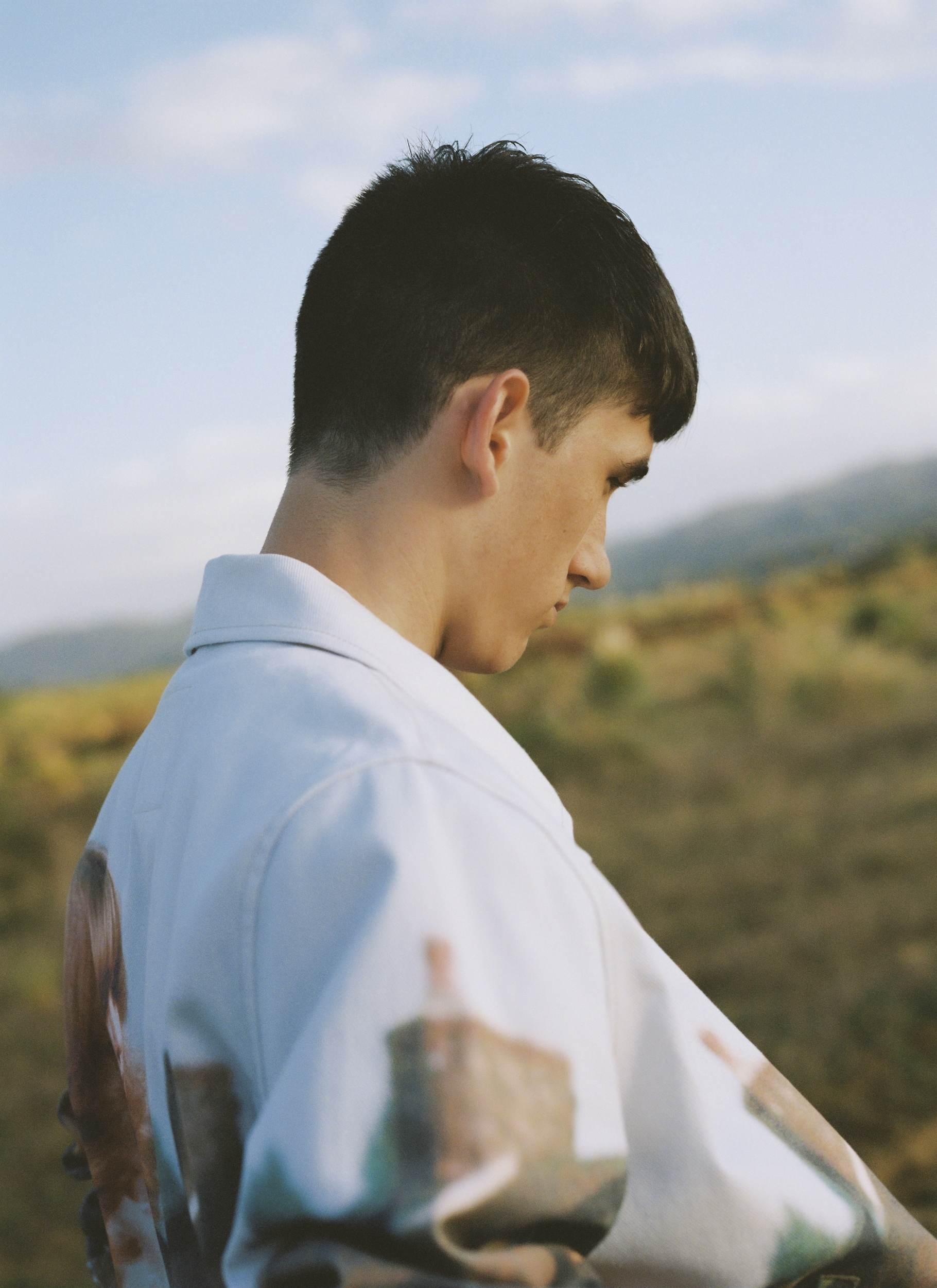 Profile portrait of a young man wearing a patterned white jacket outdoors under a clear sky.