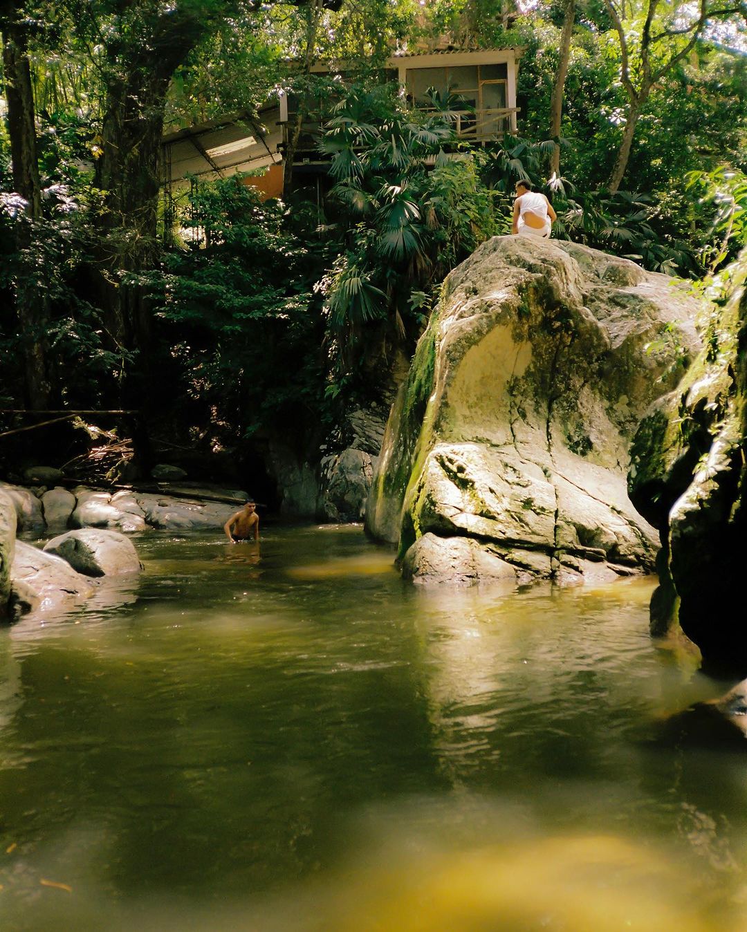 Two people in a lush green forest by a stream, one on a rock and the other in the water, surrounded by trees and a cabin.