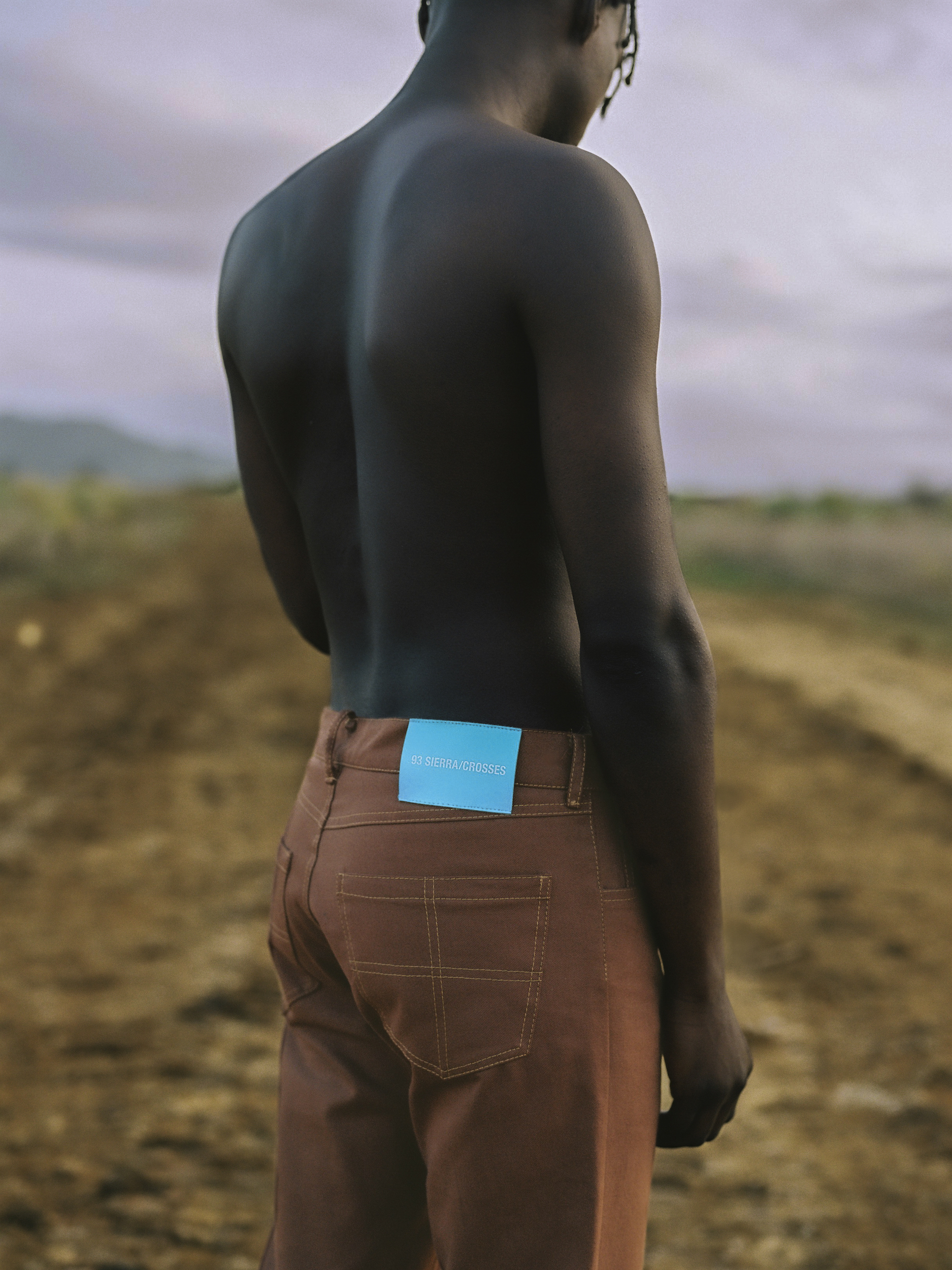 Back view of shirtless African American man wearing brown pants standing outdoors on a rural road against a cloudy sky at dusk.